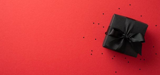 Zwart vrijdagconcept. Bovenaanzicht foto van stijlvolle zwarte geschenkdoos met lint strik en confetti op geïsoleerde rode achtergrond met copyspace - Foto, afbeelding