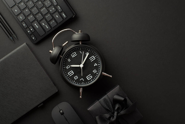 Black friday concept. Top view photo of black gift box with ribbon bow alarm clock planner pencils computer mouse and keyboard on isolated black background with copyspace - Fotó, kép