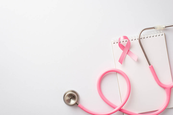 Koncept povědomí o rakovině prsu. Horní pohled fotografie růžové hedvábné stuhy stetoskop a notebook na izolovaném bílém pozadí - Fotografie, Obrázek
