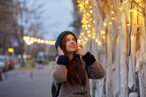Мрійлива біла жінка посміхається на вечірній вулиці. Міські вогні. Зимовий повсякденний одяг. Святкове місто
 - Фото, зображення