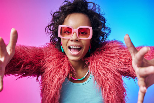 Happy African woman in trendy clothes stretching out hands and smiling against colorful background - Photo, Image
