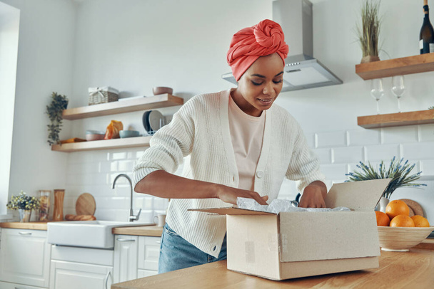 Beautiful African woman unpacking box while standing at the domestic kitchen - Foto, Bild