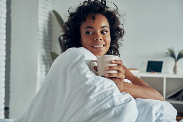 Attractive African woman holding coffee cup and smiling while enjoying lazy time in bed - 写真・画像
