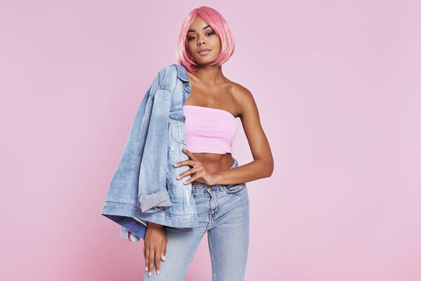 Beautiful African woman carrying denim shirt on shoulder while standing against pink background - Фото, изображение
