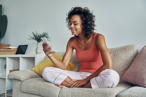 Attractive young African woman looking at her smart phone while relaxing on the couch at home - Foto, Imagem