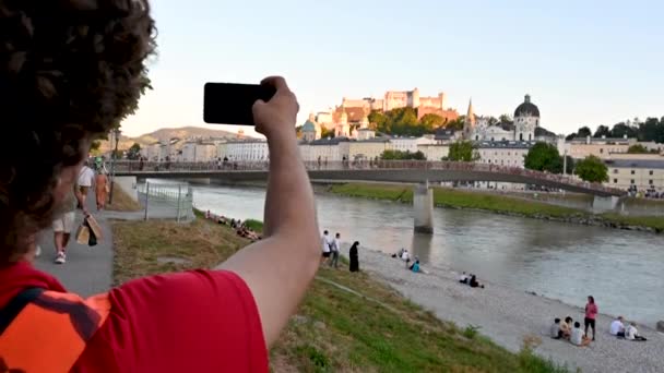 オーストリアのザルツブルク、 2022年8月。素敵な昼間の映像-若いですcaucasian男とともにパドック橋で写真を撮る古い町で支配的な砦でぼやけた背景. - 映像、動画