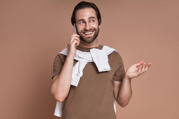 Happy young man talking on mobile phone and gesturing while standing against brown background - Foto, Imagem