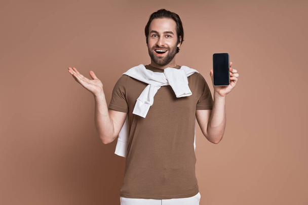 Happy young man holding smart phone and gesturing while standing against brown background - Фото, зображення