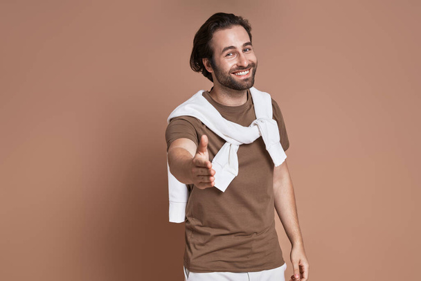 Happy young man stretching out hand for shaking while standing against brown background - Fotografie, Obrázek