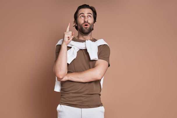 Surprised young man pointing up while standing against brown background - Foto, Imagen