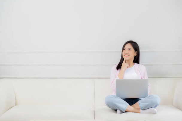 Young asian woman working on laptop computer sitting on sofa in the living room at home, female is freelance sitting on couch using notebook and doubt about decision, one person, lifestyle concept. - Photo, image