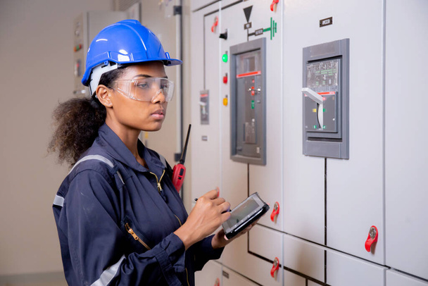 Electrical young asian woman engineer examining maintenance cabinet system electric and using tablet in control room at industrial factory, technician or electrician inspection power distribution. - Foto, Imagen