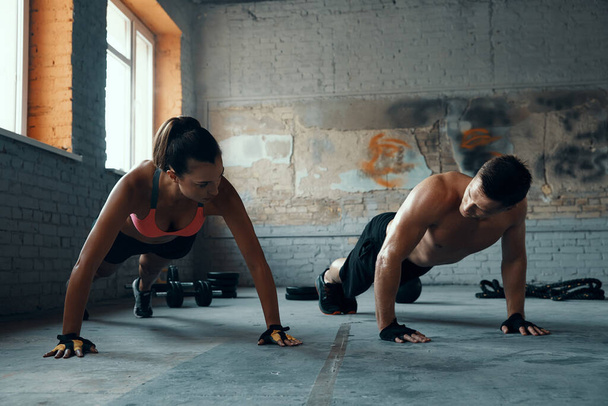 Young fit couple looking at each other while doing push-ups in gym together - Foto, imagen