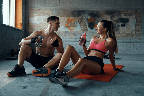 Happy young couple drinking water and talking while sitting on exercise mats in gym - Foto, Bild