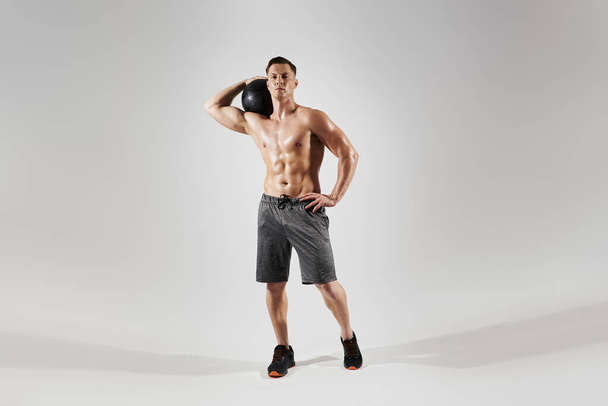 Young fit man carrying medicine ball on shoulder while standing against white background - Fotografie, Obrázek
