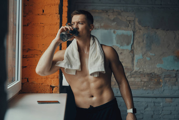 Handsome muscular man drinking water after training while standing near the window in gym - Foto, Imagem