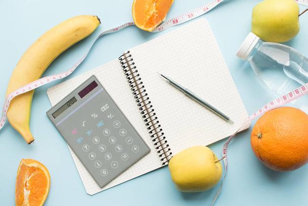 Empty paper notebook, calculator with numbers 1200, pen, bottle of water, measuring tape and fruits on blue background. Healthy eating concept - calculate daily nutrition intake. Top view. - Photo, image