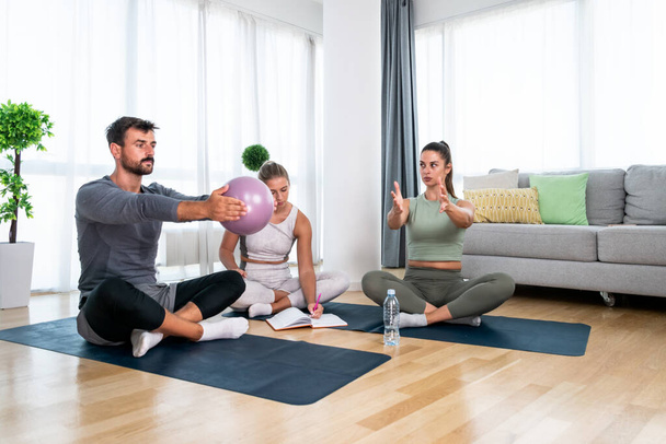 Young woman fitness and yoga instructor helping couple in their home for workout exercises. Female coach working with clients in their apartment. Husband and wife paying physical therapist for sport. - Foto, Bild