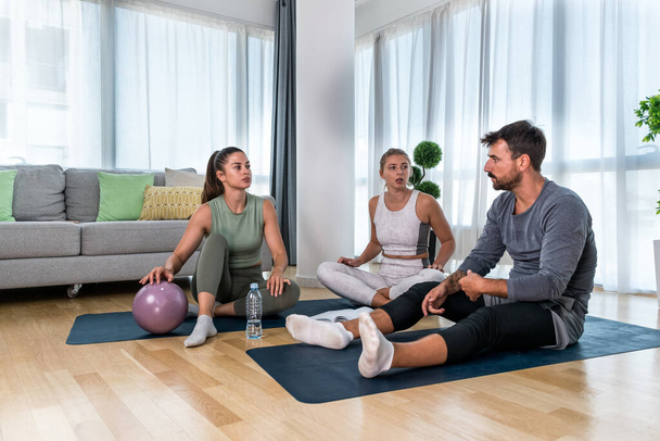 Young woman fitness and yoga instructor helping couple in their home for workout exercises. Female coach working with clients in their apartment. Husband and wife paying physical therapist for sport. - Φωτογραφία, εικόνα