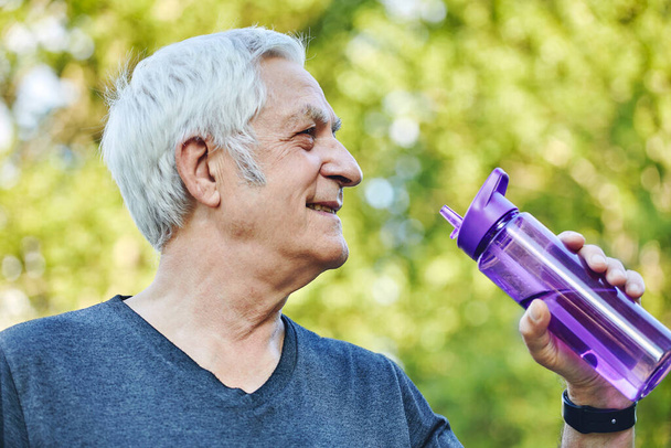 Close up image attractive mature man holds plastic reusable bottle drinking still mineral water during morning work out or stroll in summer park, caring about health, relieving thirst - Fotografie, Obrázek