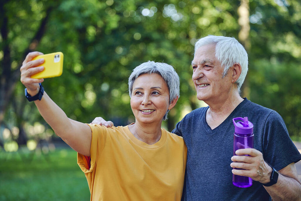 Old sportive couple staring at smart phone cam take selfie picture enjoy modern wireless tech usage while stroll in sportswear in summer park. Carefree retired life, lifestyle using technology concept - Zdjęcie, obraz