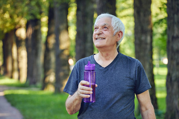 Older man in sportswear holds reusable plastic bottle standing or jogging in summer park, caring about health, enjoy morning work out feels happy. Healthy sportive lifestyle of retirees concept - Fotografie, Obrázek
