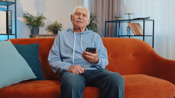 Старший дедушка, сидящий на диване, пользуется улыбкой мобильного телефона в современной квартире. Пожилой человек отправляет сообщения с контентом на смартфон в социальных сетях онлайн, смотрит расслабляющий фильм - Фото, изображение