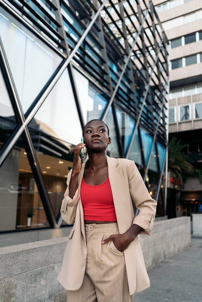 Elegant young african american woman with her hand in the pocket with short hair walking on the city looking away while talking with her phone. - Fotó, kép