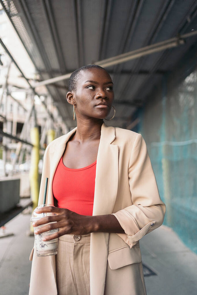 Vertical image of an elegant young african american woman holding a milkshake standing under scaffolding in a construction site looking to a side. - Фото, изображение