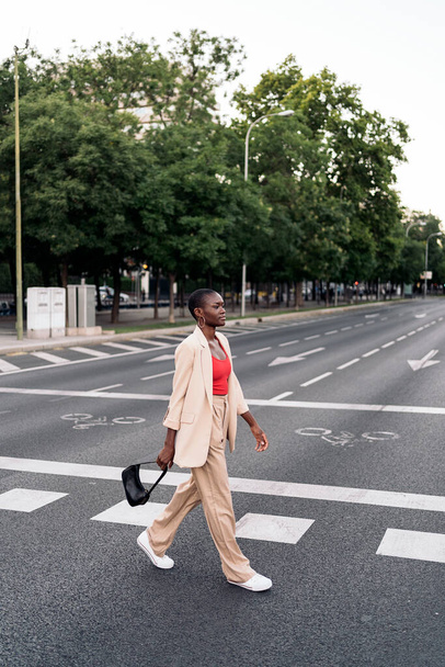 Vista completa de una elegante mujer adulta afroamericana joven con el pelo corto sosteniendo su bolso mirando hacia adelante cruzando la calle en la ciudad. - Foto, Imagen