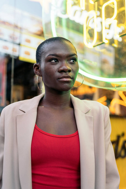 Portrait of a stylish african american adult woman standing and looking away with neon sign behind in the city - Photo, Image