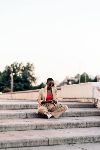 Elegant young afro american woman wearing a fashionable beige trousers and jacket set using phone outdoors. - Photo, image