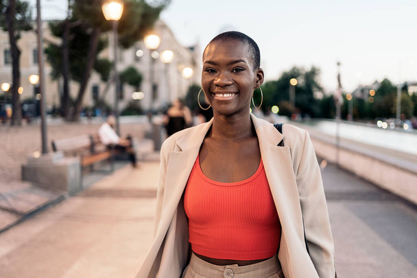 Front view of a fashionable african american young adult woman laughing while walking in the city at sunset - Valokuva, kuva