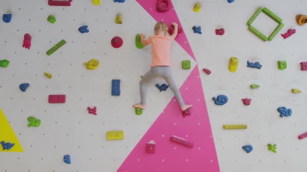 Moedig kind klein blond meisje klimt op kunstmatige muur in rotsklimmen sportschool oefening. Jeugd en extreem sportconcept. - Video