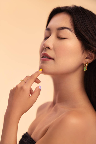 Young Asian woman with bare shoulders closing eyes and touching chin during skin care routine against beige background - Foto, Imagem