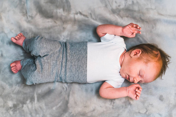 An infant baby boy with cute brown hair, laying, sleeping on his back on a grey blanket with his arms raised up. High quality photo - Photo, Image