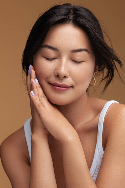 Positive Asian female with dark hair clasping hands near cheek and closing eyes during skin care routine against brown background - Фото, зображення