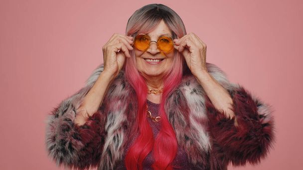 Playful happy stylish elderly woman wearing yellow sunglasses blinking eye looking at camera with toothy smile, winking and flirting expressing optimism. Senior mature grandmother on pink background - Photo, Image