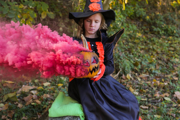 Girl dressed as witch for Halloween with pumpkin in magical smoke in the forest . - Foto, Imagem