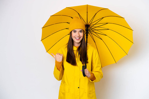 製品を提示するために側面を指して白い背景に隔離レインプルーフコートと傘を持つ若いブラジル人女性 - 写真・画像