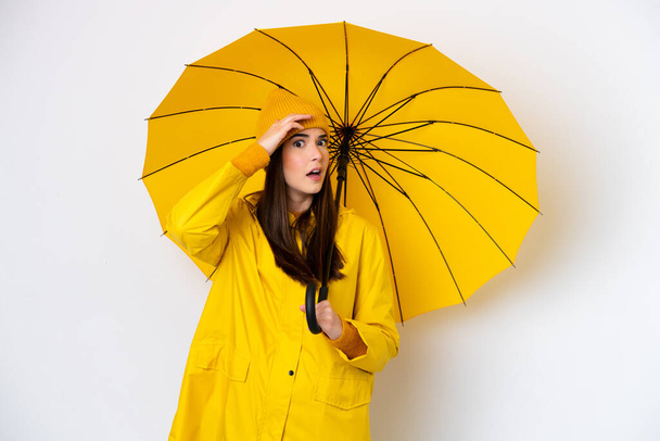 Молодая бразильская женщина в непромокаемом пальто и зонтике на белом фоне делает неожиданный жест, глядя в сторону - Фото, изображение