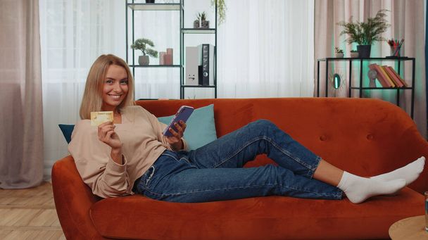 Happy shopaholic consumer young woman sitting at modern home apartment indoors making online order food delivery, payment with credit plastic bank card. Discount sale on internet. Adult girl on couch - Zdjęcie, obraz