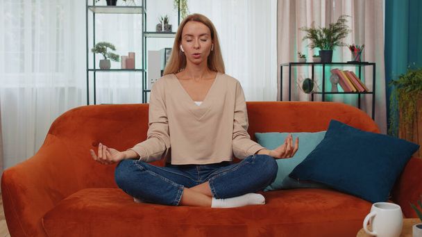 Keep calm down, relax, inner balance. Adult girl breathes deeply with mudra gesture, eyes closed meditating with concentrated thoughts, peaceful mind. Young woman sit at home in living room on couch - Photo, Image