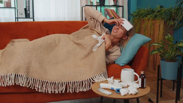 Adult ill girl suffering from cold or allergy lying on sofa in living room at home. Sick young woman with towel on head blows sneezes wipes snot into napkin. Coronavirus quarantine pandemic. Lifestyle - Photo, Image
