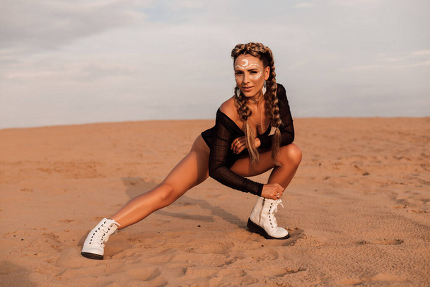 The woman has creative makeup, braided hair in braids. She is wearing a black swimsuit and shod in white boots. A girl poses sitting - Foto, Imagen