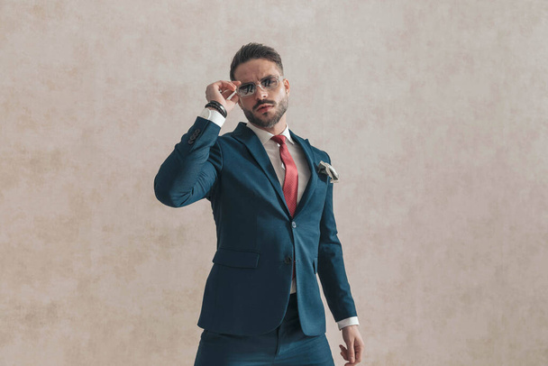 koele elegante man dragen stijlvolle outfit met zakdoek aanpassen zonnebril in de voorkant van beige achtergrond, staan en poseren in de studio - Foto, afbeelding