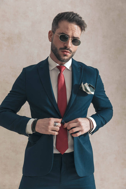 elegant bearded man with sunglasses wearing red tie and unbuttoning navy blue suit, posing in front of beige background in studio - Photo, Image