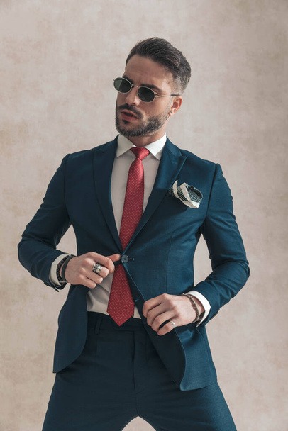 sexy unshaved businessman with glasses looking away while fixing navy blue suit, wearing red tie and handkerchief in front of beige background, posing in studio - Photo, Image
