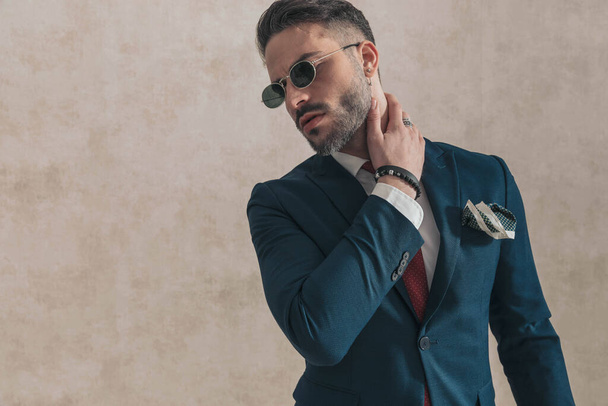 portrait of attractive businessman in suit with sunglasses holding hand behind neck and looking to side while confidently posing on beige background - Fotoğraf, Görsel