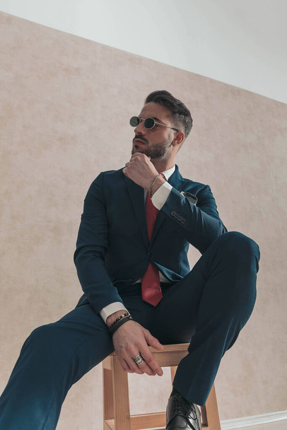 cooler nachdenklicher Mann im eleganten Anzug mit roter Krawatte und Taschentuch, der Kinn und Denken berührt, während er zur Seite schaut und auf einem Holzstuhl sitzt - Foto, Bild